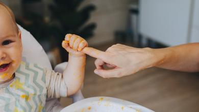 
		Baby with porridge on his face and hands holds onto a parent's fingers
	